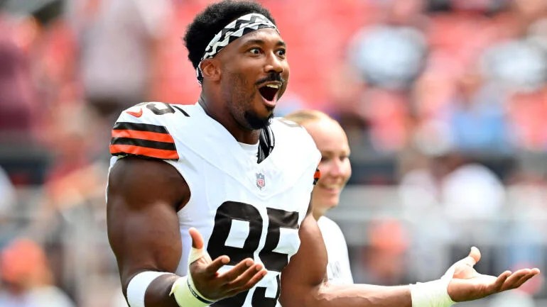 A football player in a white jersey with orange and black stripes, wearing a headband, expressing surprise and joy on the field.