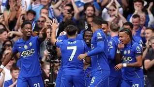 A group of soccer players in blue jerseys celebrating a goal, with fans cheering in the background.